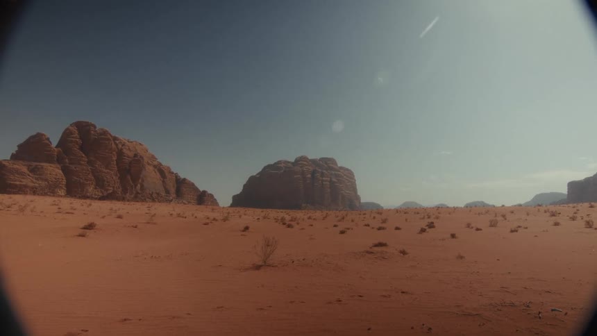 A wide, still shot captures an expansive and arid red desert landscape with towering rock formations under a bright, sunny sky