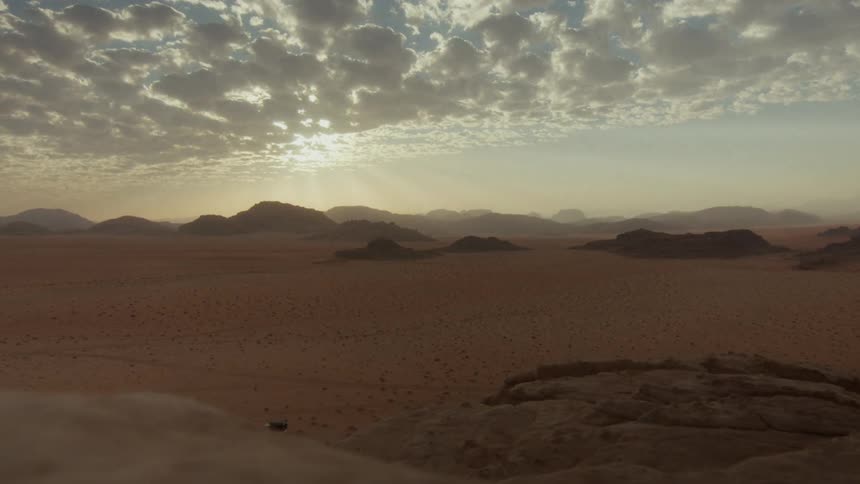 A lone white vehicle drives across the vast, scenic Wadi Rum desert landscape at sunrise under a cloudy sky
