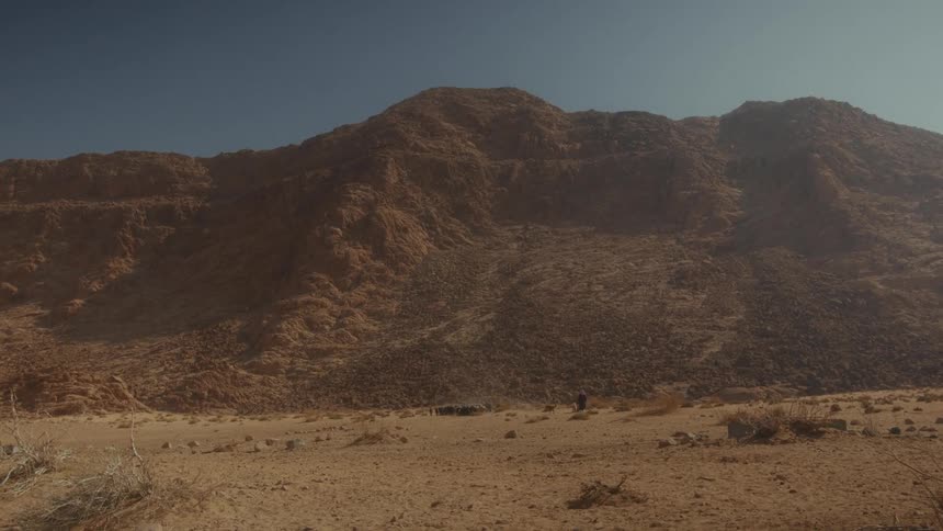 A lone shepherd tends to his flock of sheep at the base of a vast, rocky mountain in the desert