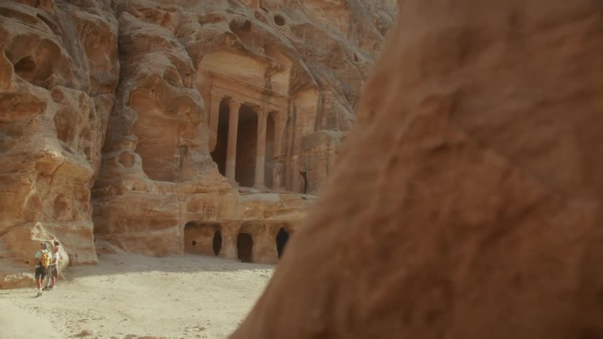 A stunning reveal shot from behind a rock formation unveils a magnificent ancient tomb carved into the sandstone cliffs of Petra as tourists look on