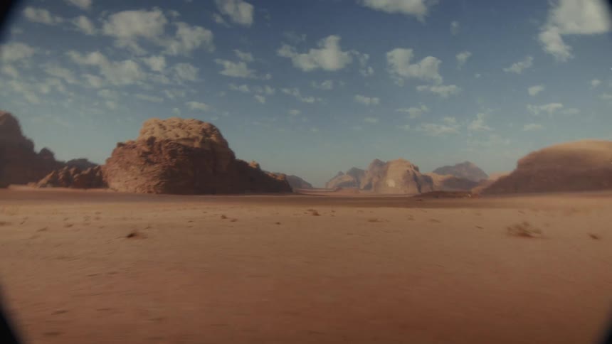 A point-of-view shot driving through a vast desert valley with large rock formations under a blue, cloudy sky