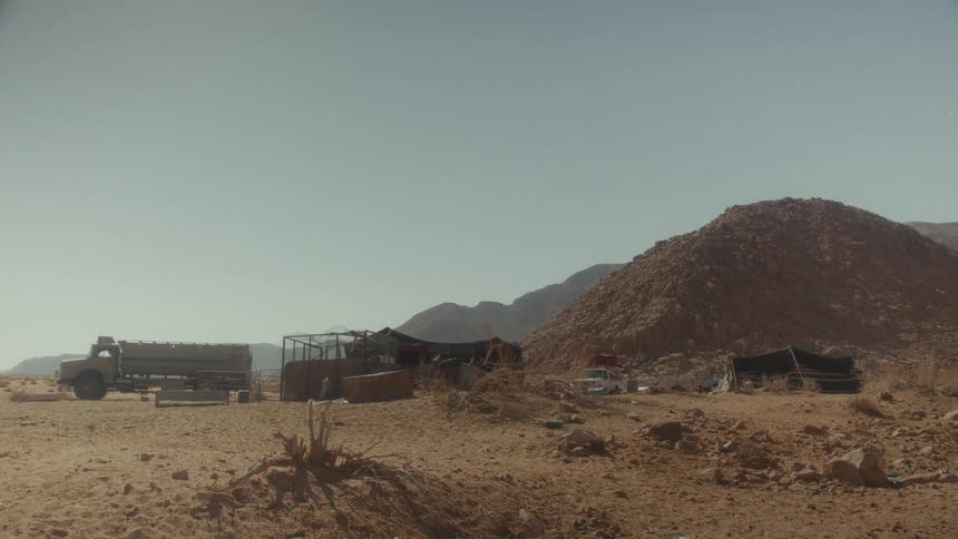 A makeshift camp with a water tanker truck sits isolated in a vast, sun-drenched desert landscape with rocky mountains in the background
