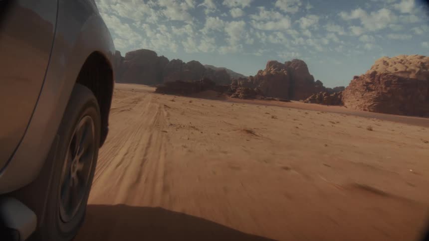 Dynamic low-angle shot of a silver SUV driving at speed across a vast sandy desert with rock formations in the background