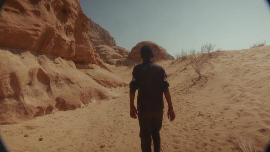 Slow motion shot from behind of a man walking through a hot, sandy desert valley surrounded by rock formations