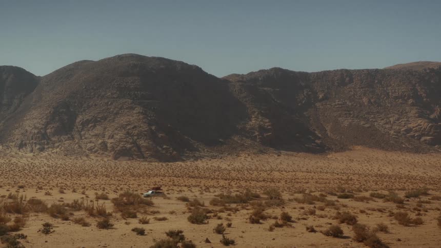 A white SUV with luggage strapped to the roof drives off-road through a vast, sunny desert with large mountains in the background