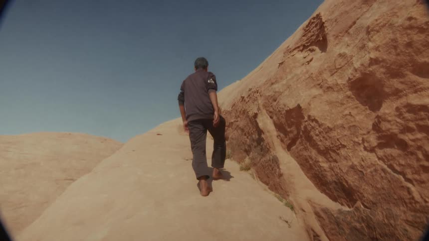 A man walks barefoot in slow motion up a massive red rock formation under a clear blue sky in the desert