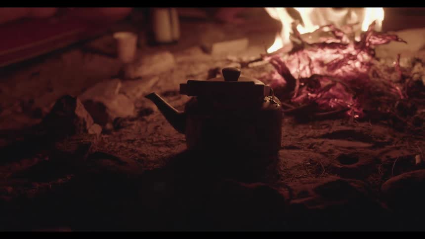 A rustic teapot is silhouetted by the warm glow of a campfire at night