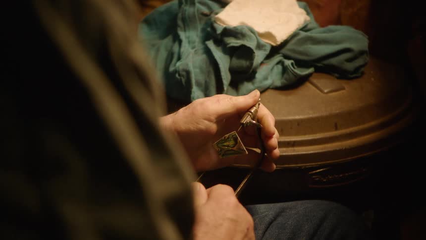 Over-the-shoulder view of a jeweler meticulously cutting a small, iridescent piece with a handsaw in a rustic workshop