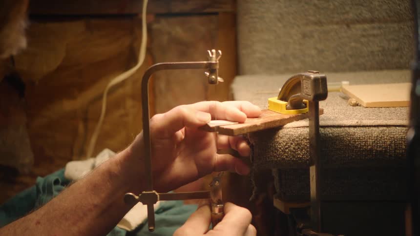 An elderly craftsman with a white beard uses a jeweler's saw for intricate work under a warm lamp in his studio