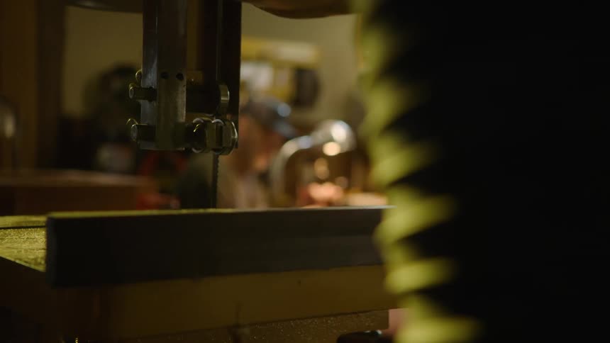 A skilled craftsman works diligently at his workbench in a dimly lit workshop as the focus shifts to a bandsaw in the foreground