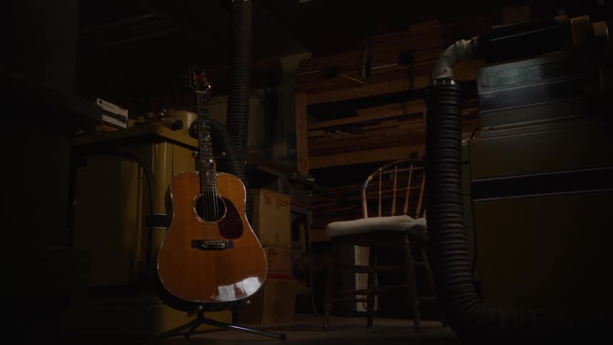 An acoustic guitar stands illuminated by a single light source in a dark woodworking workshop