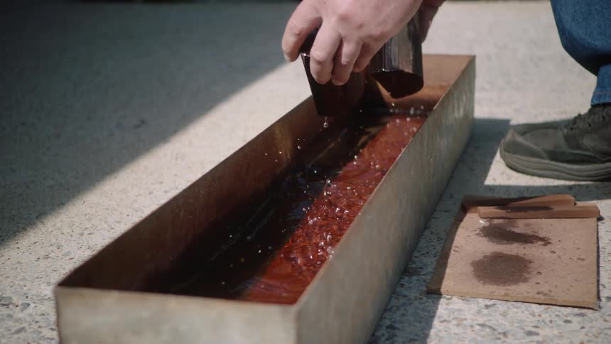 In slow motion, a craftsman dips a dark wooden block into a metal trough filled with reddish wood stain