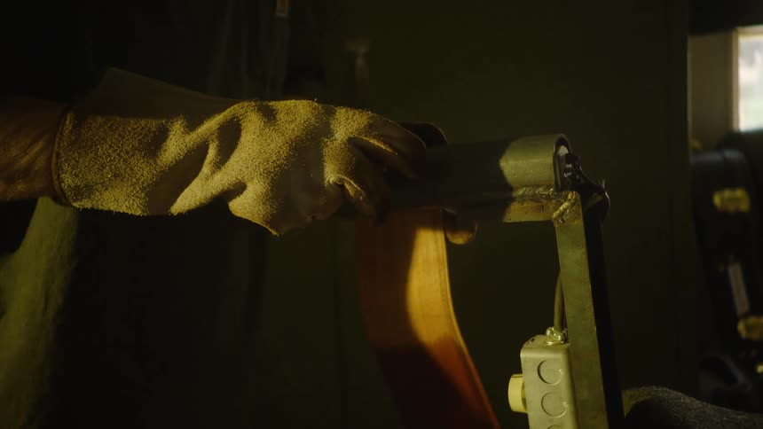 A craftsman wearing gloves carefully sands a curved piece of wood on a belt sander in a dimly lit workshop