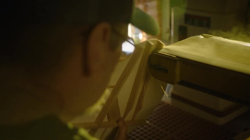 A craftsman carefully sands a wooden creation on a belt sander in a dimly lit workshop, with sawdust flying in slow motion