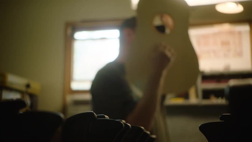 A luthier holds up the unfinished wooden body of an acoustic guitar, inspecting his craftsmanship in a warm workshop