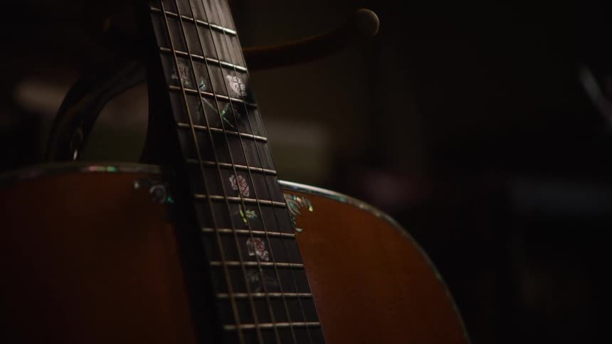 A slow focus pull reveals intricate floral inlays on the fretboard of an acoustic guitar in a dimly lit setting