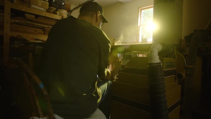A woodworker meticulously sands a piece of wood on a large sanding machine in his sunlit workshop, with sawdust filling the air