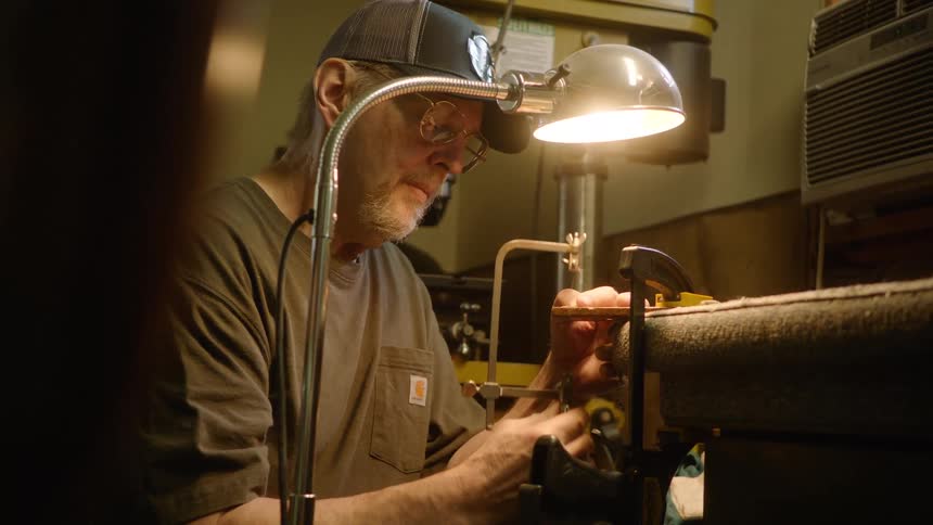 A skilled artisan meticulously saws a small part in his workshop, illuminated by a single, bright lamp