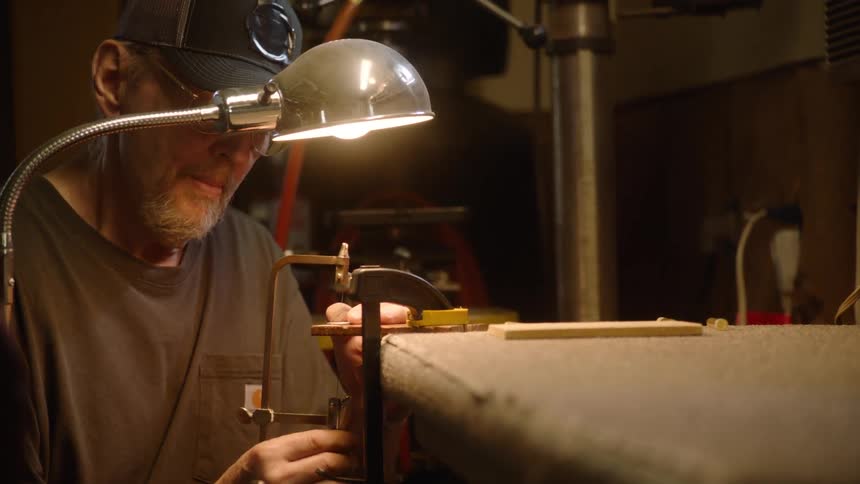 A focused craftsman meticulously uses a coping saw to cut a small piece of wood under a single lamp in his workshop