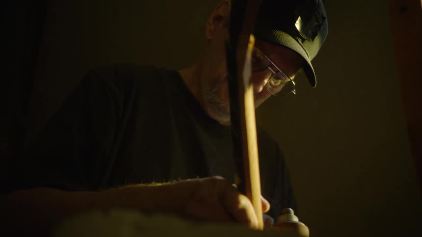 A focused craftsman wearing a hat with a light carefully sands the neck of a guitar in a dimly lit workshop