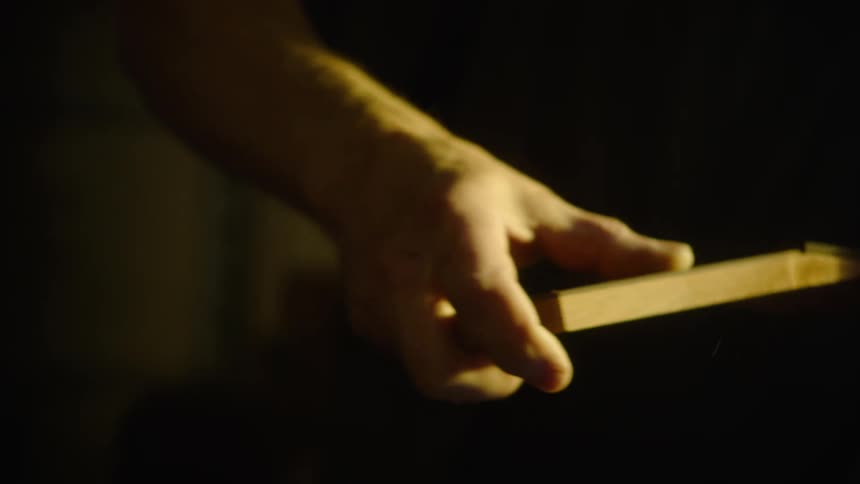 A luthier carefully sands and shapes the neck of a guitar on a spindle sander in a dimly lit workshop, with sawdust flying in slow motion