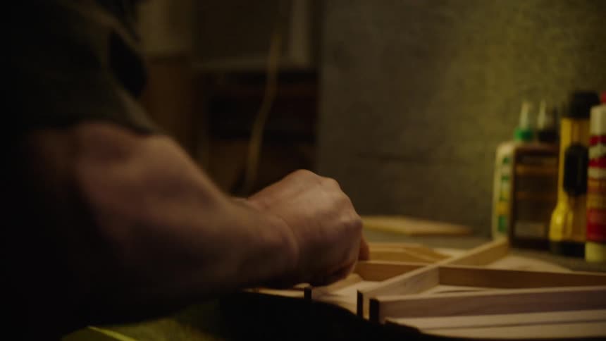 A craftsman in his dimly lit workshop meticulously assembles the intricate wooden bracing for an acoustic guitar