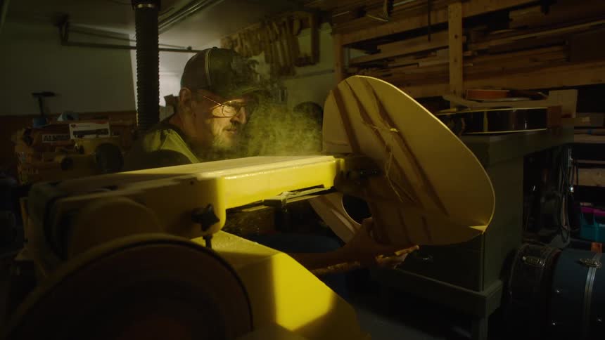 A master craftsman carefully sands the wooden body of a guitar in his workshop, creating a cloud of smoke and sawdust in slow motion
