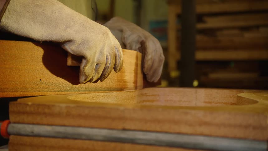 A craftsman carefully bends a piece of heated wood into a guitar-shaped mold in a dimly lit workshop