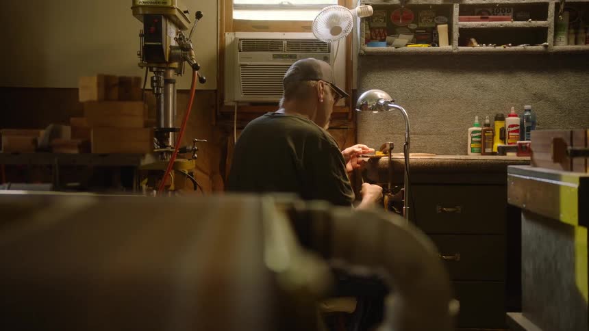 A craftsman sits at his workbench, illuminated by a lamp as he meticulously works on a detailed project in his workshop