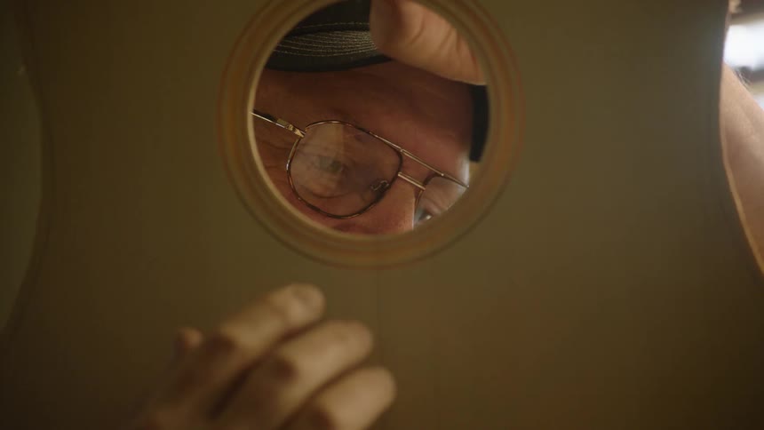 A luthier with glasses carefully inspects the interior of a handcrafted wooden guitar through the soundhole in his workshop