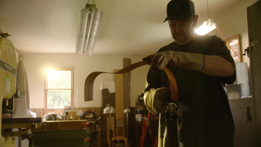 An artisan in protective gloves carefully works with a long, thin strip of wood in his sunlit workshop