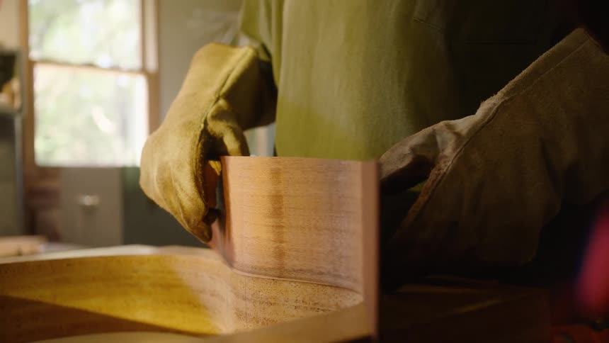 In slow motion, a luthier wearing protective gloves carefully shapes a piece of heated wood into a guitar mold in a workshop