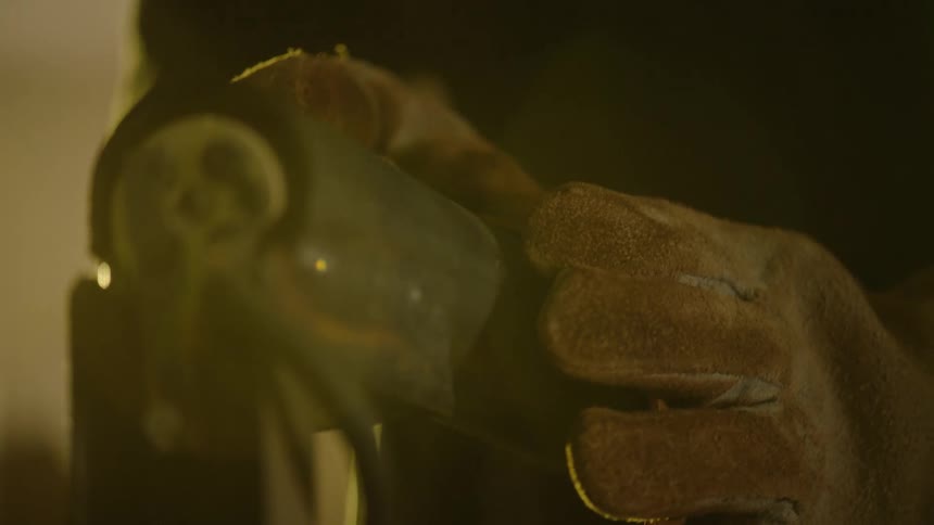 A close-up slow-motion shot of a worker's gloved hands grinding a piece of metal on a belt sander in a workshop, causing sparks to fly