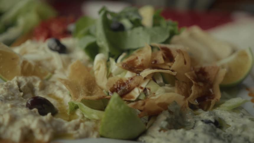 A delicious platter of Mediterranean meze, including hummus, tzatziki, salad, and fried pita bread, is revealed in a slow-motion close-up