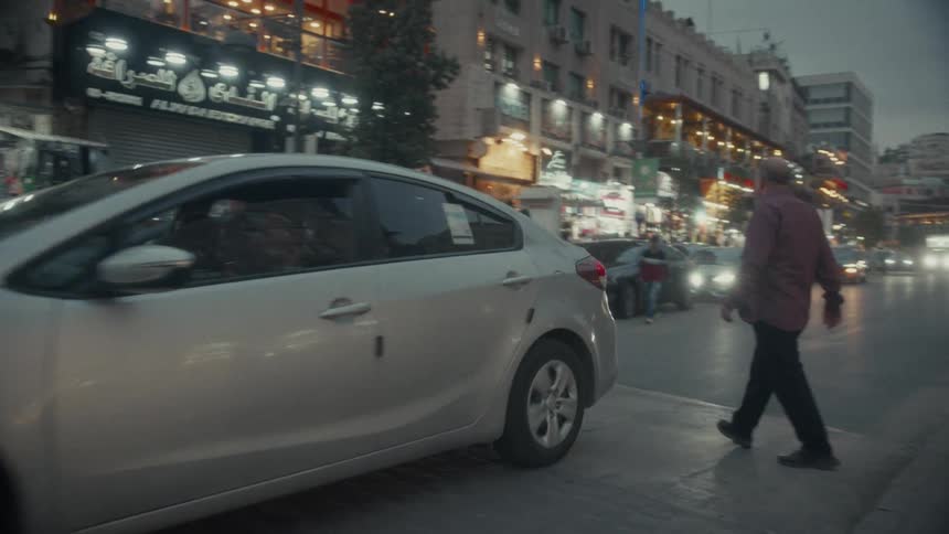 A man walks down a busy city street at dusk in the Middle East as cars drive by brightly lit storefronts