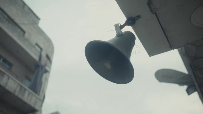 A low-angle shot of a public address system speaker hanging from a building under a gloomy, overcast sky