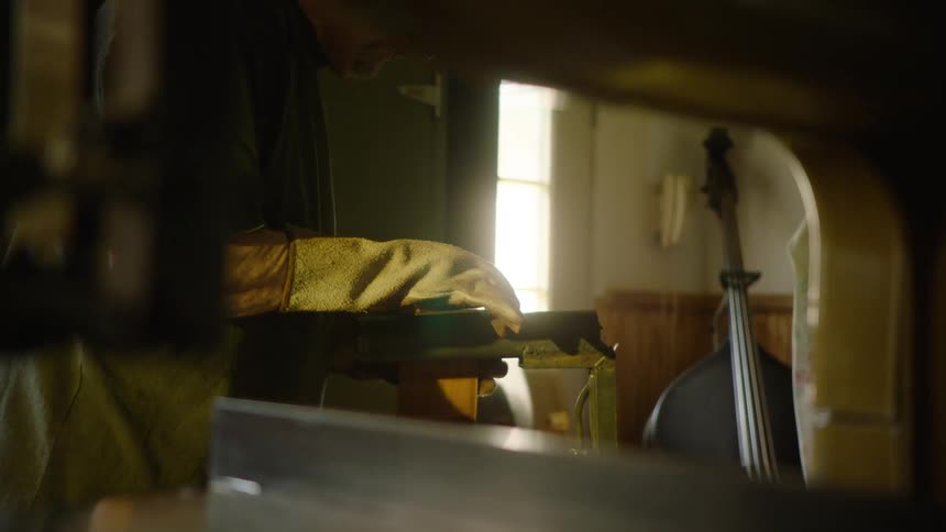 A luthier in protective gloves carefully uses a band saw to cut and shape the headstock of a musical instrument in his workshop
