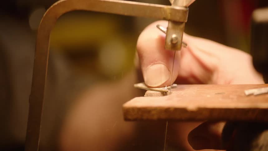 A craftsman meticulously saws a detailed initial from a small metal sheet in a dimly lit workshop