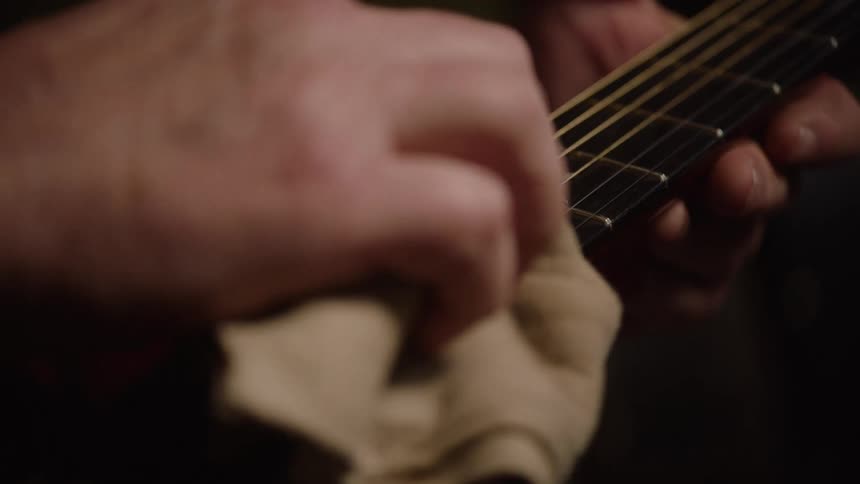 A musician carefully cleans and polishes the fretboard and strings of an acoustic guitar in slow motion