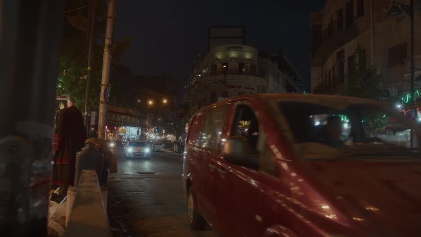 A red van drives down a bustling city street in Amman, Jordan at night as pedestrians walk past illuminated shops