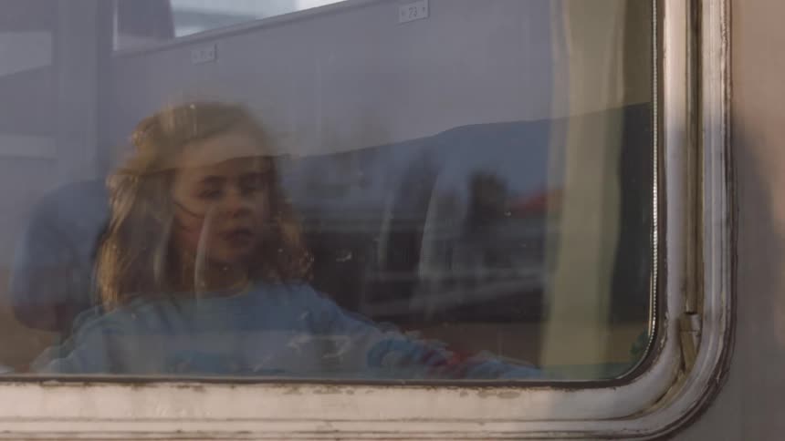 A pensive young girl with curly hair looks out the window of a moving train, her reflection visible in the glass