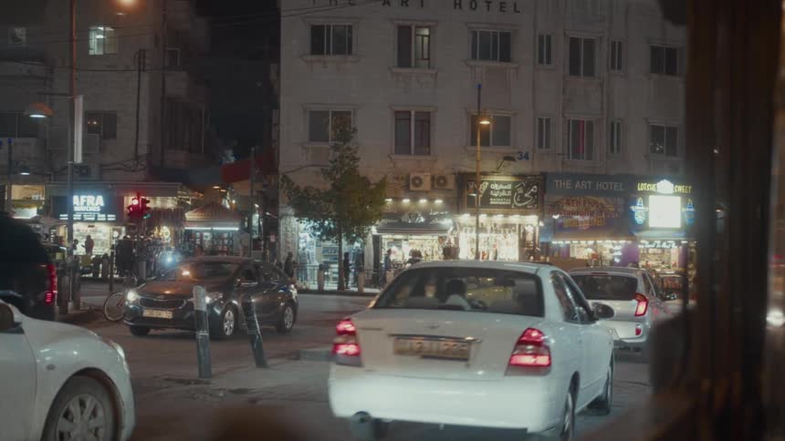 Cars navigate a busy street intersection in a Middle Eastern city at night, passing brightly lit storefronts and 'The Art Hotel'