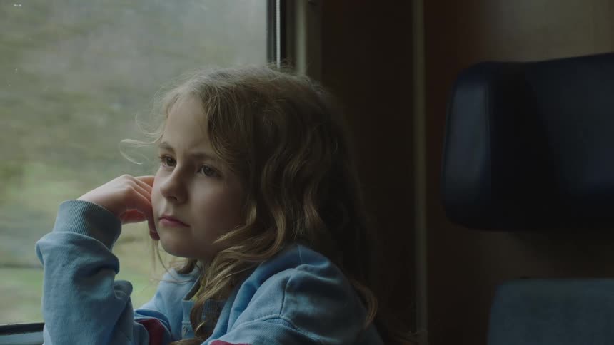 A pensive young girl with curly blonde hair rests her head on her hand as she looks out the window of a moving train