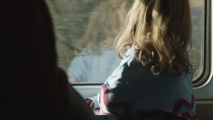 A pensive young girl with long blonde hair gazes out the window of a moving train, her reflection watching back