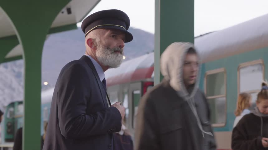 An elderly train conductor signals departure with a whistle on a crowded station platform
