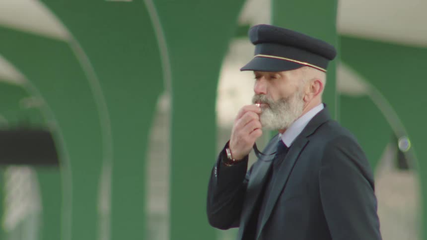 A senior train conductor in uniform blows his whistle to signal the departure of the train from the station platform