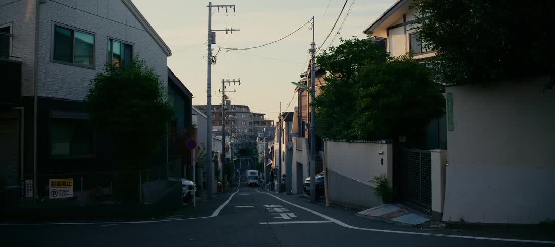 A quiet, sloping residential street in a Japanese neighborhood is bathed in the warm light of dusk