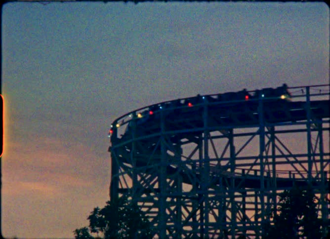 Vintage 8mm footage shows a wooden roller coaster with colorful lights moving along the track against a dusky sky