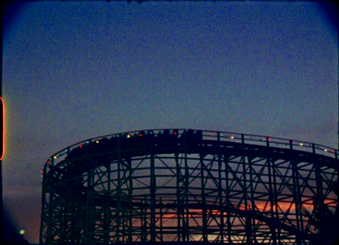 A wooden roller coaster is silhouetted against the evening sky as a train of cars moves along the track in this vintage 8mm footage