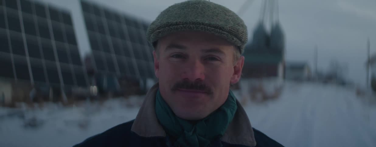 Portrait of a friendly man with a mustache and a flat cap smiling warmly in a snowy landscape with solar panels in the background